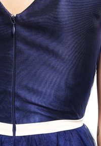 Tmavě modré šaty s texturovanou látkou, V-výstřihem a viditelným zipem na zadní straně, s kontrastním bílým páskem.