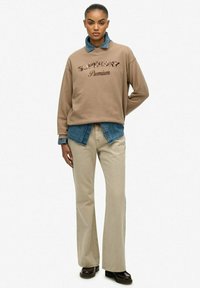 Brązowy sweter z napisem "Superdry Premium", nałożony na dżinsową koszulę. Połączony z beżowymi spodniami i brązowym obuwiem, całość ma swobodny charakter.