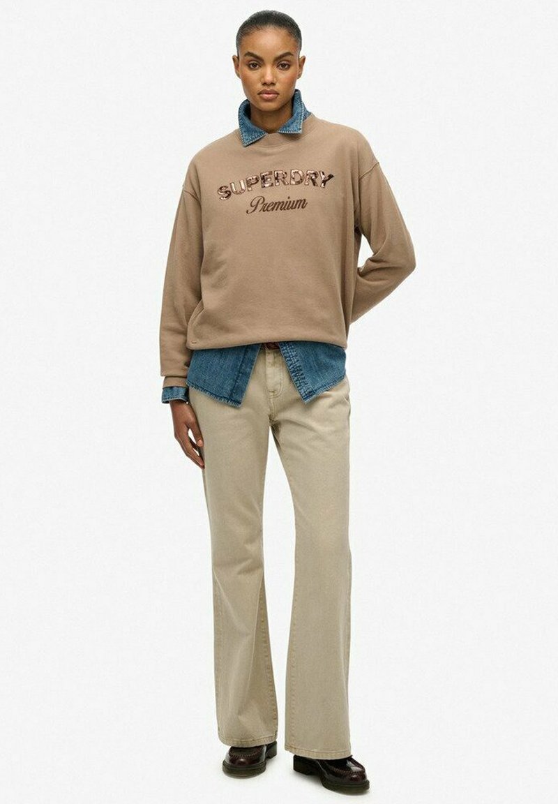 Brązowy sweter z napisem "Superdry Premium", nałożony na dżinsową koszulę. Połączony z beżowymi spodniami i brązowym obuwiem, całość ma swobodny charakter.