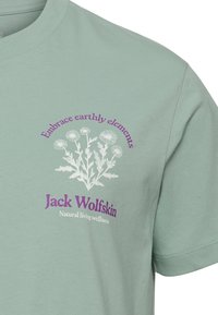 Light green cotton t-shirt featuring white floral graphic and purple text: "Embrace earthly elements" and "Jack Wolfskin." Casual crew neck design.