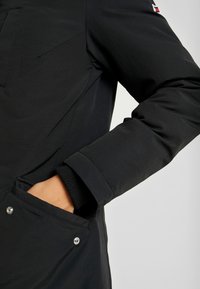 Personne portant une veste noire avec la main dans une poche avant à boutons-pression, logo de la marque visible sur le haut du bras sur un fond uni.