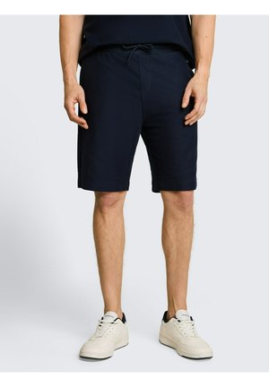 Mann trägt schwarze Shorts mit Kordelzug und weiße Sneakers, steht vor einem einfachen hellen Hintergrund.