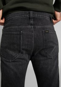 Bakifrån av en person som bär svarta denimjeans med bakfickor och en Lee-märkesetikett på den högra fickan, händerna nedstuckna i midjan.