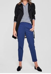 Czarny trencz założony na niebieską bluzkę w paski, niebieskie dopasowane spodnie i czarne wsuwane buty. Strój wyróżnia się podwiniętymi mankietami i ozdobnymi guzikami.