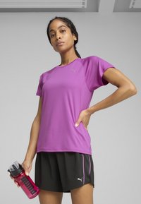 Lilla træningsskjorte med korte ærmer, kombineret med sorte shorts med pink accenter, holdt sammen med en gennemsigtig sportsflaske.