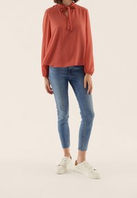 Blusa corallo a maniche lunghe con dettaglio a chiave e nodo, abbinata a jeans blu slim e sneakers bianche con dettagli dorati.