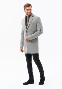 Cappotto grigio di lana con un solo bottone, due tasche laterali e revers a punta. Indossato sopra un maglione a righe verdi e abbinato a jeans neri e stivali.
