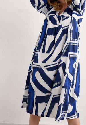 Vrouw die een losse jurk tot op de knie draagt met opvallende abstracte blauwe en witte geometrische patronen, terwijl ze haar armen boven haar hoofd heft.
