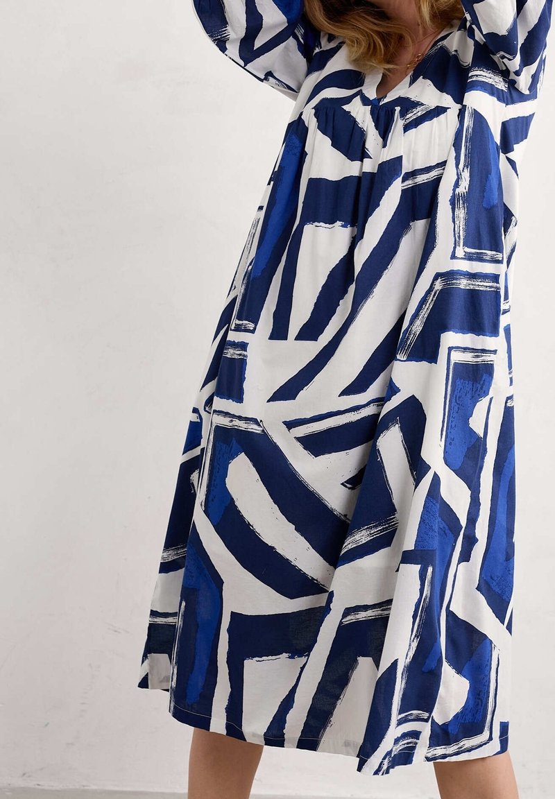 Femme portant une robe ample et mi-longue avec des motifs géométriques abstraits bleu et blanc audacieux, levant les bras au-dessus de la tête.