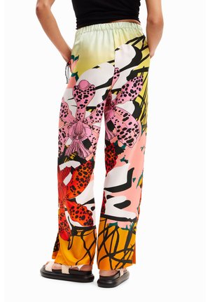 Pantalones de pierna ancha con un gran estampado floral en rosa, rojo, blanco y negro, combinados con una blusa negra y sandalias blancas, mostrados desde atrás.