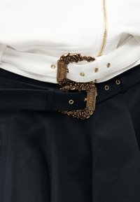 Valged ja mustad nahkvööd rikkalike kuldsete pandlatega, millel on graveering "Versace Jeans Couture", üle valge jaki ja mustade pükste.