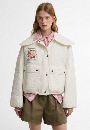 Jonge vrouw draagt een witte doorgestikte Barbour Farm Rio jas met bloemenpatch, rood gestreept overhemd en olijfgroene shorts, staand tegen een witte achtergrond.