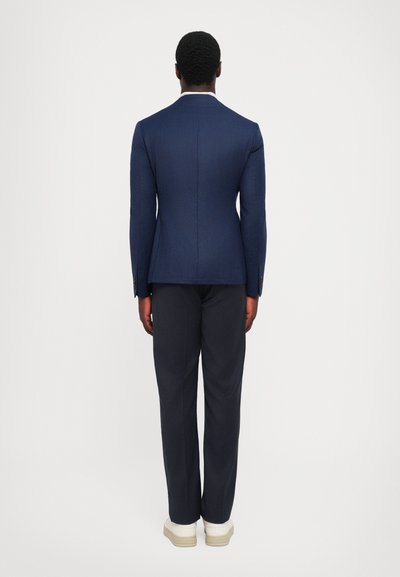 Blazer bleumarin și pantaloni închiși, croi atent, țesătură texturată, vent unic la spate, asortați cu adidași albi.