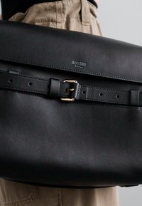 Geanta satchel din piele neagră Moschino, cu un curea cu cataramă, ținută de o persoană îmbrăcată în pantaloni bej și un top negru, pe un fundal simplu.