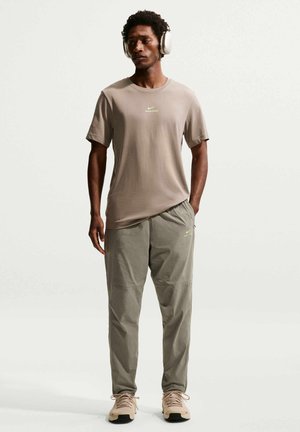 Homme portant un t-shirt Nike Pro beige, un pantalon Nike Pro vert olive, des baskets beiges et un casque audio blanc, debout devant un fond blanc.