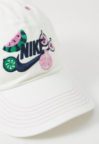Λευκό καπέλο μπέιζμπολ με πολύχρωμα κεντήματα που σχετίζονται με φρούτα και εμφανές ναυτικό λογότυπο Nike. Περιλαμβάνει ροζ ραφές και καμπυλωτό γείσο.