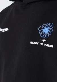 Tissu noir avec un graphisme abstrait de fleur bleue au-dessus du texte blanc "READY TO WEAR" et un petit symbole d'étoile blanche en dessous.