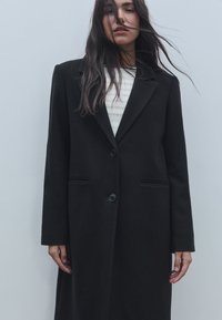 Musta villakangastakki, jossa on räätälöity muotoilu, varustettuna lovikolarilla, kahdella napilla ja sivutaskuilla. Suora siluetti, jossa on sileä rakenne.