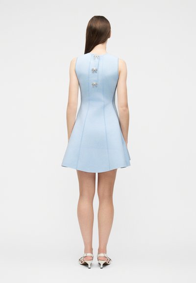 Femme portant une mini-robe bleu clair sans manches avec trois petits nœuds dans le dos, assortie à des sandales blanches à talons, debout de dos.