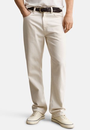 Man wearing off-white straight-leg pants, white sneakers, a white shirt tucked in, and a dark braided belt, standing with hands relaxed.