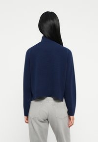 Persona con lunghi capelli neri che indossa un maglione blu navy e jeans grigio chiaro, in piedi con la schiena rivolta verso la fotocamera.