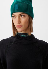 Teealfarbene Strickmütze mit geripptem Textur und einem rechteckigen Logo-Patch. Das Modell trägt einen schwarzen Pullover mit hohem Kragen und sichtbarem Reißverschluss.