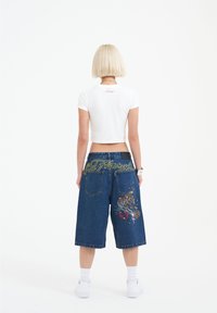 Blue denim cropped shorts with large embroidered design on the back, featuring colorful patterns. Paired with a white fitted short-sleeve top.