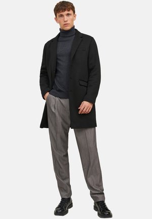 Junger Mann, der einen schwarzen Mantel, einen dunkelgrauen Rollkragenpullover, graue Nadelstreifenhosen und schwarze Lederstiefel trägt, steht mit einer Hand in der Tasche.