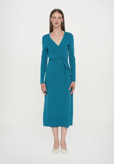 Blågrön, långärmad omlottklänning med v-ringning, markerad midja och knytband. Tyget har en slät textur, klänningen når till mitten av vaden och matchas med beiget högklackat.