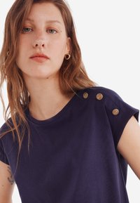 Mørkeblå T-shirt med korte ærmer, rund halsudskæring og fire dekorative guldknapper på venstre skulder. Glat stofstruktur.