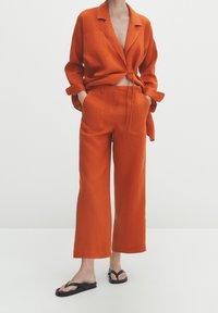Oranje linnen twee-delig set met een cropped blazer met strik aan de voorkant en brede pijpen. Draag het met zwarte slippers, waarbij de gladde stoftextuur te zien is.
