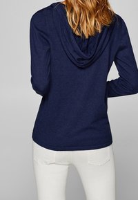 Marineblaue Kapuzenjacke mit lockerer Passform, ausgestattet mit einer drapierten Kapuze, strukturiertem Stoff und gerippten Bündchen, kombiniert mit weißen Jeans.