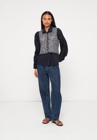 Γκρίζα μοτίβα αμάνικη μπλούζα πάνω από ένα σκούρο μακρυμάνικο πουκάμισο, συνδυασμένη με φαρδιά μπλε τζιν και σαγιονάρες, σε λευκό φόντο.