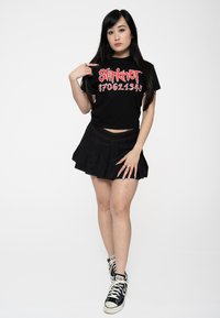 Black crop top with red Slipknot text, paired with a black pleated mini skirt. Worn with black high-top sneakers. Neutral background.