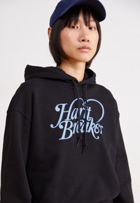 Svart hettegenser med et frontgrafikk i lyseblå tekst "Heart Breaker." Har snor i hetten og en avslappet passform. Bært med en blå caps.