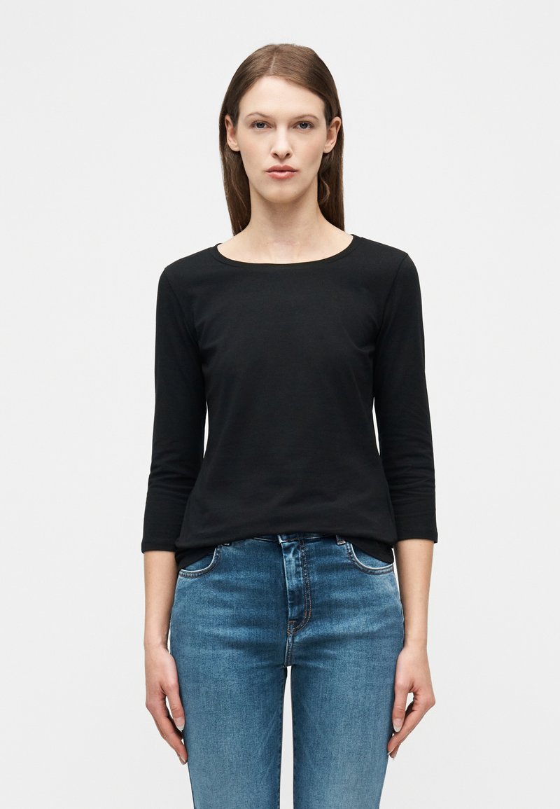 Γυναίκα με μακριά καστανά μαλλιά, που φοράει μαύρο τοπ με μανίκια τριών τετάρτων και μπλε τζιν, στέκεται μπροστά σε λευκό φόντο.