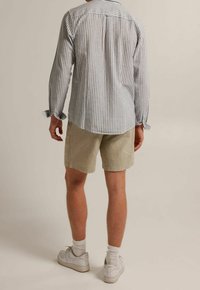 Hombre vestido con pantalones cortos beige, una camisa de rayas azul y blanca de manga larga, calcetines blancos y zapatillas Nike Air blancas, de pie y mirando hacia atrás.