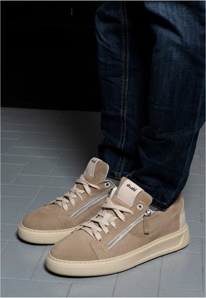 Beige suede sneakers with side zippers and laces worn with dark blue jeans on gray tiled floor.