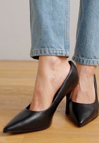 Scarpe nere in pelle con tacco alto, punta affusolata e lati aperti, abbinate a pantaloni in denim azzurro, evidenziando una texture liscia.