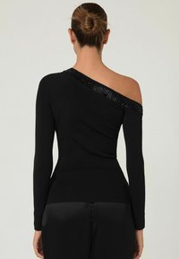 Žena s vlasmi v drdole, oblečená v čiernom topu s jedným ramenom a dlhým rukávom, ktorý má ozdoby po odhalenom ramene, a čiernych nohaviciach.