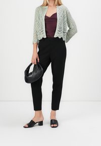 Helle grüne Strickjacke über einem bordeauxfarbenen Oberteil, kombiniert mit schwarzen, maßgeschneiderten Hosen und schwarzen Blockabsatz-Slides. Trägt eine schwarze Tasche.