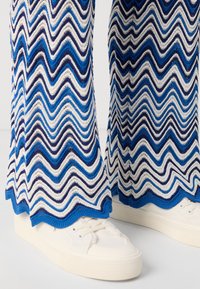 Striped knit pants feature a zigzag pattern in blue and white, with a scalloped hem. Paired with white platform sneakers.