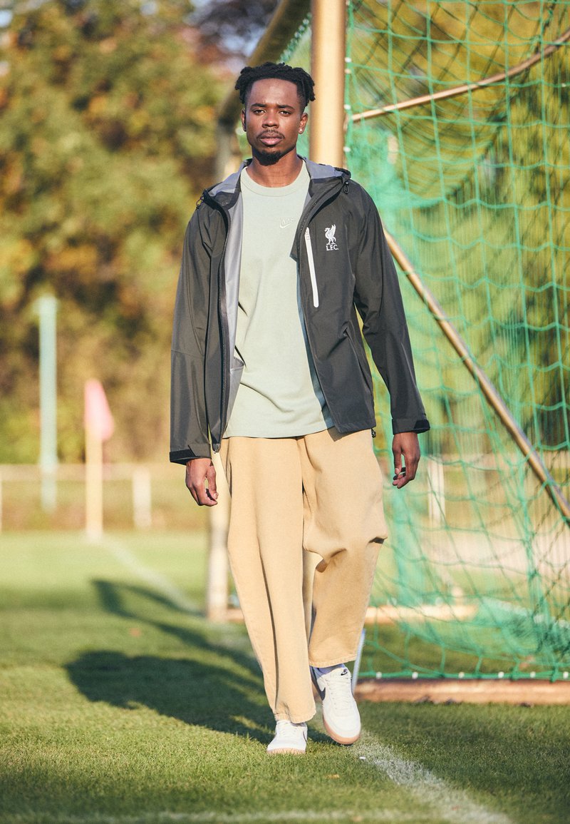 Schwarze leichte Regenjacke mit grauen Akzenten, grünes T-Shirt, beigefarbene Hose und weiße Sneakers, stehend auf Gras in der Nähe eines Torpfostens.