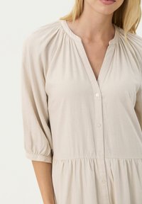 Rochie bej cu nasturi, cu mâneci bufante 3/4, textură moale, decolteu plisat și design cu volane la margine.