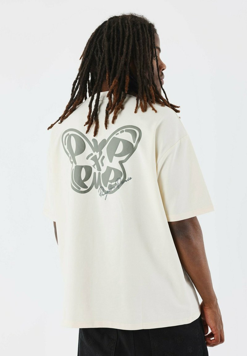 Camiseta oversized color crema con un gran gráfico de mariposa gris y texto en la parte de atrás. Fabricada con una tela suave y lisa, con un corte relajado.