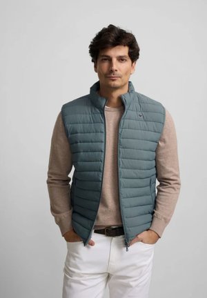 Hombre de cabello oscuro con chaleco acolchado azul sobre un suéter beige y pantalones blancos, de pie con las manos en los bolsillos frente a un fondo liso.