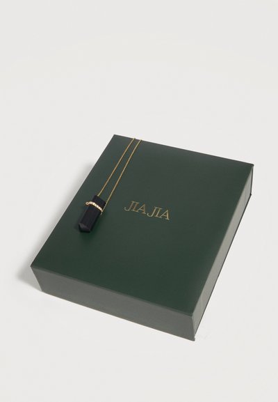 Χρυσή αλυσίδα με μαύρο κρύσταλλο μενταγιόν και μικρό χρυσό δαχτυλίδι, που ξεκουράζεται σε σκούρο πράσινο κουτί με χρυσό ανάγλυφο όνομα μάρκας JALA.