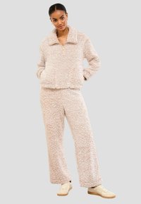 Pahuljasta, svijetlosmeđa loungewear set s jaknom s patentnim zatvaračem i širokim hlačama. Mekana tekstura s labavim krojem, uparena sa svijetlim tenisicama.
