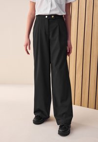 Pantalon noir à jambes larges et taille haute, doté de boutons dorés et d'une texture lisse, associé à des chaussures noires.