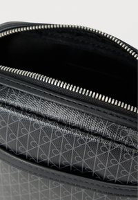 Μαύρη τσάντα με φερμουάρ, με υφή εξωτερικής επιφάνειας που παρουσιάζει γεωμετρικό μοτίβο. Το εσωτερικό είναι επενδεδυμένο με μαύρο ύφασμα και έχει ορατό μηχανισμό φερμουάρ.